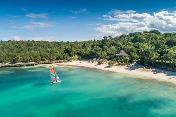 Tropical beach with sailboat and lush trees.