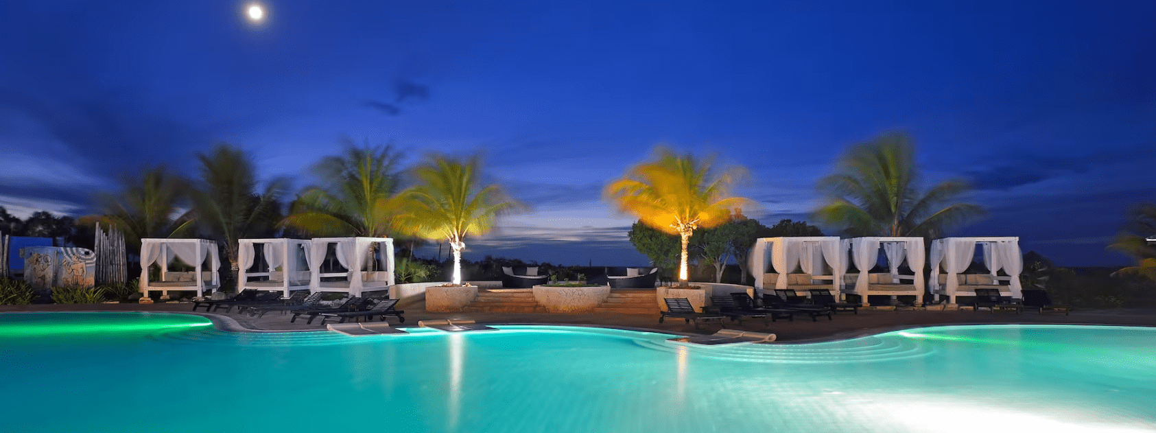 Nighttime pool with cabanas and palm trees.