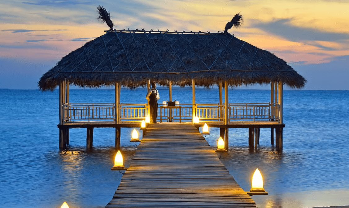 Lit path to thatched hut over water.