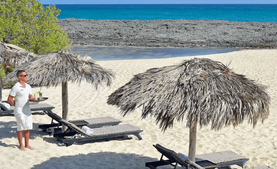 Man serving drinks on a beach with palm umbrellas.