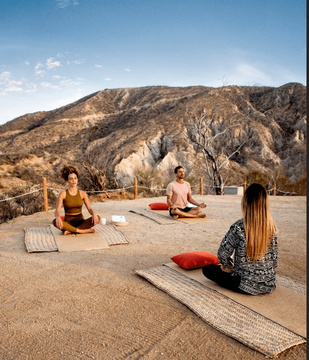 Three people meditate outdoors in desert.