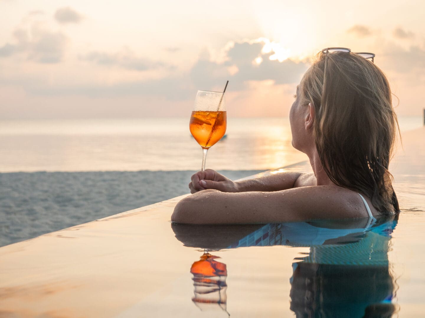 Woman in pool at sunset holding a drink.