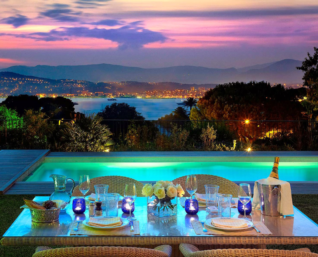 A dinner table set in front of a pool with a view of the sea.