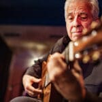 Mature male Guitarist on the stage with classic guitar. Grey hair. He is wearing all black. Stage interior of Theatre during performance at night.